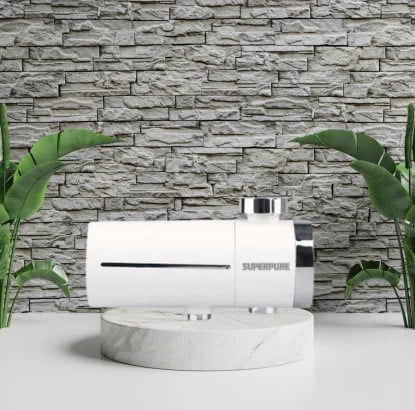 The SUPERPURE TAPURE Tap-Mounted Water Filter is showcased against a modern stone wall backdrop, flanked by fresh green plants, emphasizing clean water directly from your tap.